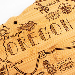 The Destination Oregon Cutting Board by Totally Bamboo, measuring 14-1/2" x 10-3/4", is a decorative piece shaped like the state of Oregon. It features a laser-engraved map highlighting places such as Maupin, Madras, and Pendleton, along with artistic illustrations of regional icons and national forests.