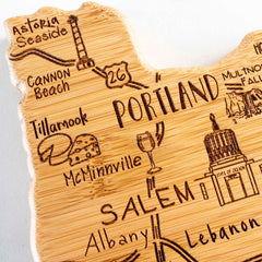 Introducing the "Destination Oregon Cutting Board" by Totally Bamboo, this 14-1/2" x 10-3/4" state-shaped wooden board is elegantly laser-engraved with a detailed map of Oregon. It showcases cities such as Portland and landmarks like Cannon Beach, adorned with charming icons like a wine glass and cheese. Both functional and decorative, it offers a quality bamboo cutting surface reminiscent of craftsmanship found in traditional pieces.