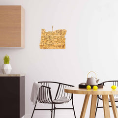 A modern kitchen features black chairs surrounding a round wooden table adorned with a teapot and yellow apples. A geometric vase containing a green plant is situated on the counter, alongside the sleek Destination Oregon Cutting Board by Totally Bamboo. On the white wall hangs a decorative wooden map of Oregon.