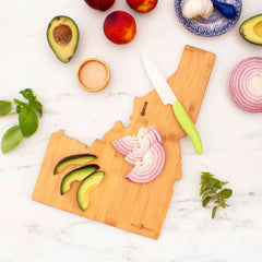 A Destination Idaho Cutting Board by Totally Bamboo, measuring 15" x 10-1/2", displays laser-engraved artwork and holds sliced red onion and avocado alongside a nearby knife. The board is surrounded by whole avocados, peaches, garlic, a bowl of salt, fresh basil, and oregano on a marble countertop.