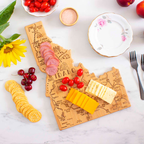 A Destination Idaho Cutting Board by Totally Bamboo, measuring 15" x 10-1/2", is artfully adorned with laser-engraved artwork and topped with sliced meats, cheese, and tomatoes. It sits amidst crackers, cherries, a sunflower, a plate, and forks on a marble surface.