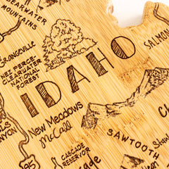 A close-up of the Destination Idaho Cutting Board by Totally Bamboo reveals the intricate details of a wooden map in the shape of Idaho. Laser-engraved artwork highlights notable places such as Nez Perce-Clearwater National Forest and New Meadows, with "Idaho" prominently showcased in stunning fashion on a 15" x 10-1/2" board.