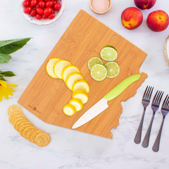 A Destination Arizona Cutting Board by Totally Bamboo intricately features the state shape, holding sliced lemons and limes. A knife with a green handle rests beside them. Surrounding the artistic display are cherry tomatoes, peaches, a sunflower, crackers, and four forks on a marble surface.