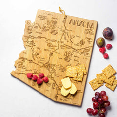 Introducing the Totally Bamboo Destination Arizona Cutting Board, measuring 13-1/2" x 11-3/4". This wooden board beautifully captures the silhouette of Arizona, featuring laser-engraved city names and landmarks. It's elegantly styled on a white background with crackers, cheese, raspberries, figs, and grapes.