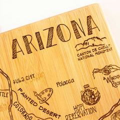 The "Destination Arizona Cutting Board" by Totally Bamboo, measuring 13-1/2" x 11-3/4", is crafted from wood and laser-engraved with a depiction of Arizona. It highlights landmarks like "Canyon de Chelly National Monument," "Painted Desert," and "Tuba City." Decorative engravings of a turtle and mountain sketch are included to capture the region's beauty.