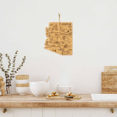 In a warm and minimalist kitchen, a laser-engraved wooden Arizona state map decorates the white wall. Below, the countertop is adorned with elegant white bowls, a jar, the "Destination Arizona" cutting board by Totally Bamboo measuring 13-1/2" x 11-3/4", and freshly baked cookies. A vase filled with eucalyptus adds a refreshing touch of greenery to complete the scene.