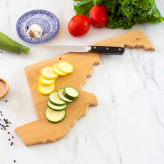 A Totally Bamboo Destination Maryland Cutting Board (18" x 9-5/8") serves as both a practical kitchen tool and stunning wall art, beautifully arranged with sliced yellow and green squash. A knife rests on the board, accompanied by a bowl of garlic, tomatoes, lettuce, and peppercorns on a marble countertop.