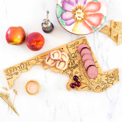 A Totally Bamboo Destination Maryland Cutting Board, measuring 18" x 9-5/8", holds sliced cheese and sausage. Nearby are crackers, two peaches, a flower-patterned plate, a small bowl of seasoning, a pepper grinder, and some grapes on a marble surface.