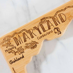 The Destination Maryland Cutting Board by Totally Bamboo, measuring 18" x 9-5/8", serves as a unique wall art piece. It features engraved Maryland landmarks such as Fort McHenry and Deep Creek Lake, set against a wavy-edged border that stands out beautifully on its marble-like backdrop.