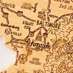 Introducing the Totally Bamboo Destination Maryland Cutting Board, measuring 18" x 9-5/8", which showcases a detailed wooden map of Maryland, USA. Ideal as wall art, it highlights key locations such as Baltimore and Annapolis, along with surrounding areas like Rockville and Gaithersburg. The board also features decorative elements like a small train and geographic details such as the Liberty Reservoir.