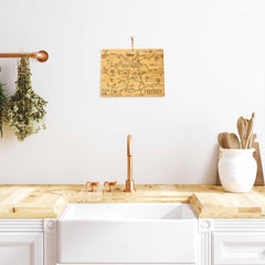 A minimalist kitchen showcases a farmhouse sink paired with a rose gold faucet. Wooden utensils sit in a holder, and herbs are suspended from a copper rail. A bamboo cutting surface is accompanied by the Totally Bamboo Destination Colorado Cutting Board, measuring 14-1/2" x 10-3/4", hanging on the white wall above the countertop.