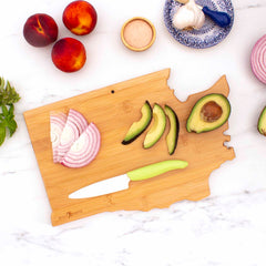 A Destination Washington Cutting Board by Totally Bamboo, measuring 15-1/2" x 10", features sliced onions, avocados, and a knife. Nearby are whole peaches, garlic cloves, and a small bowl of salt. The state-shaped cutting board elegantly rests on a white marble surface.