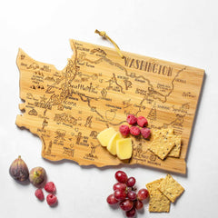 The Totally Bamboo Destination Washington Cutting Board, measuring 15-1/2" x 10", is finely crafted in the shape of Washington state and adorned with intricate laser-engraved artwork showcasing various cities and landmarks. This wooden cutting board elegantly displays slices of cheese, crackers, raspberries, and figs. Nearby, grapes and additional crackers are thoughtfully arranged for an extra touch of enjoyment.