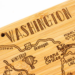 This is the Destination Washington Cutting Board by Totally Bamboo, measuring 15-1/2" x 10". This state-shaped cutting board features a laser-engraved, stylized map of Washington with highlighted regions such as "Okanogan," "Winthrop," and "Grand Coulee." It is adorned with illustrations like a hot air balloon and mountain on a strong bamboo surface.