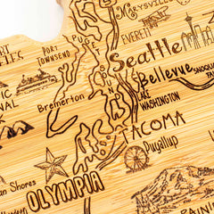 The Totally Bamboo Destination Washington Cutting Board, measuring 15-1/2" x 10", is a wooden carving featuring a laser-engraved artwork of a stylized map of Washington state. It includes city names such as Seattle, Tacoma, and Olympia and highlights natural landmarks like Puget Sound and Lake Washington. Decorative details include a compass rose and pine tree motif.