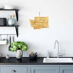 A modern kitchen with gray cabinets and a black countertop features a white sink and silver faucet. Decorative items and potted plants adorn the counter and shelves, complemented by the Totally Bamboo Destination Washington Cutting Board, a laser-engraved artwork of Washington state, hanging on the white wall.