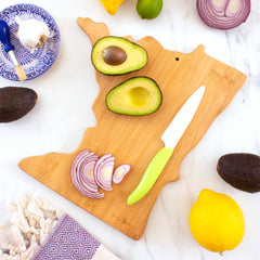 A Totally Bamboo Destination Minnesota Cutting Board, measuring 13-1/4" x 11-3/4", is adorned with sliced avocado and red onion, complemented by a green-handled knife. In a nearby bowl on a marble surface, a lemon, lime, and garlic are accompanied by a fringed towel.