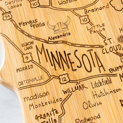 Introducing the Totally Bamboo Destination Minnesota Cutting Board, measuring 13-1/4" x 11-3/4": a state-shaped board with a bamboo cutting surface, engraved with a detailed map of Minnesota. It highlights towns such as Alexandria, Morris, and St. Cloud and features decorative details like a Viking helmet illustration near Alexandria.