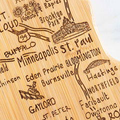 Introducing the Destination Minnesota Cutting Board by Totally Bamboo, a beautifully carved wooden map highlighting Minnesota's charm. This 13-1/4" x 11-3/4" state-shaped board features intricately etched illustrations of notable cities like St. Cloud, Minneapolis, St. Paul, and Hastings, along with depictions of local buildings and natural scenery.