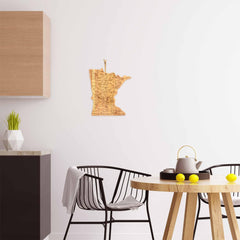 In a contemporary kitchen, a round wooden table with black chairs creates a cozy ambiance. A teapot and yellow cups are arranged on the tabletop. On the nearby white wall, a Destination Minnesota Cutting Board by Totally Bamboo is displayed, adding character alongside a cabinet and a small plant for an inviting touch.