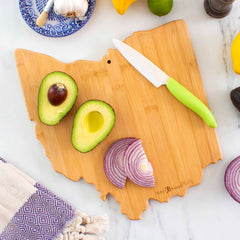 A Totally Bamboo Destination Ohio Cutting Board, measuring 12" x 13-1/2", rests on a marble surface. Sliced avocado and red onion adorn the board, while a white knife with a green handle sits nearby. A blue-patterned plate holds a garlic bulb and lemon, complemented by a fringed towel for added charm.