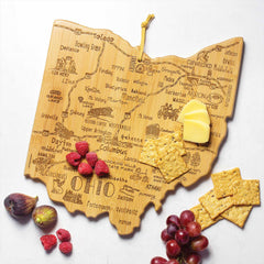 The Destination Ohio Cutting Board by Totally Bamboo, measuring 12" x 13-1/2", is shaped like the state of Ohio and features intricate laser-engraved artwork showcasing city names and landmarks. It elegantly displays an artistic arrangement of cheese, crackers, grapes, figs, and raspberries for a perfect presentation.