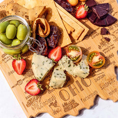 The Destination Ohio Cutting Board from Totally Bamboo, measuring 12" x 13-1/2", beautifully presents an enticing selection of snacks, including herb-infused cheese, strawberries, and a jar of green olives. Accompanied by pita chips, dates, cherry tomatoes, and pieces of dark chocolate, the laser-engraved city names on the board add a distinctive flair to this vibrant arrangement.