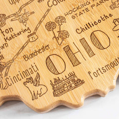 The Destination Ohio Cutting Board by Totally Bamboo, measuring 12" x 13-1/2", serves as both a state-shaped cutting board and a carved wooden map of Ohio, highlighting cities like Cincinnati and Chillicothe. It is adorned with playful illustrations such as a winged unicorn and a steamboat, while decorative flowers embellish the region marked Hocking State Park on its distinctive bamboo surface.