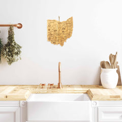 A modern kitchen showcases a white sink with a wooden countertop, adorned with a copper faucet. The space is enhanced by the Totally Bamboo Destination Ohio Cutting Board, measuring 12" x 13-1/2", which adds to the décor. Dry herbs hang nearby, while a map of Ohio decorates the wall. Wooden utensils are neatly arranged in a holder on the right, creating a minimal and stylish ambiance.