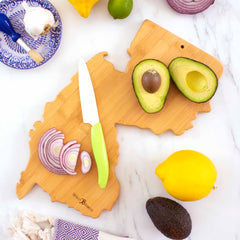 A Destination New Jersey Cutting Board by Totally Bamboo, measuring 8-5/8" x 16-5/8" and featuring laser-engraved artwork of New Jersey, displays a halved avocado and sliced red onion. Nearby, a knife with a green handle rests alongside a whole avocado, lemon, garlic, lime, and a blue-patterned plate on a sleek marble surface.