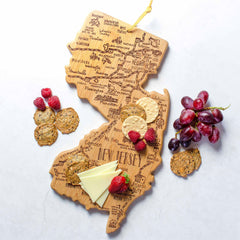 The Destination New Jersey Cutting Board by Totally Bamboo, measuring 8-5/8" x 16-5/8", elegantly highlights the state of New Jersey with laser-engraved town names. The board is surrounded by a tasteful arrangement of crackers, sliced cheese, raspberries, and grapes on a spotless white surface.