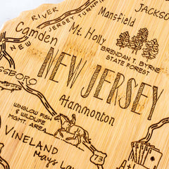 The Destination New Jersey Cutting Board by Totally Bamboo, measuring 8-5/8" x 16-5/8", features a laser-engraved wooden map of New Jersey with intricate illustrations and text that highlight locations like the Jersey Turnpike, Camden, Mt. Holly, and Vineland. Adorned with drawings of a horse, trees, and a fish and wildlife area, this cutting board is both artful and informative.