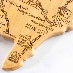 The Destination New Jersey Cutting Board by Totally Bamboo, measuring 8-5/8" x 16-5/8", showcases a close-up of a state-shaped bamboo surface. It features laser-engraved locations in New Jersey, such as Cape May, Ocean City, and Atlantic City. The board is decorated with illustrations that emphasize the areas and attractions, all presented in a rustic style.