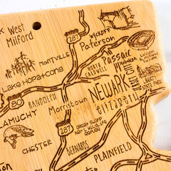 The Destination New Jersey Cutting Board, measuring 8-5/8" x 16-5/8" from Totally Bamboo, is a bamboo cutting board shaped like the state and showcases a laser-engraved map of northern New Jersey with major highways and cities such as Newark and Paterson. It also features decorative illustrations, including a fish, bridge, and compass that enhance its unique artwork.