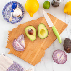 On the Totally Bamboo Destination Georgia Cutting Board, 11-3/4" x 13-1/4", there is a halved avocado and sliced red onion. Nearby are a knife with a green handle, garlic, lemons, a lime, a whole avocado, and a blue patterned plate.