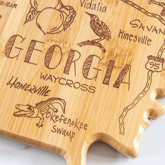 The Totally Bamboo Destination Georgia Cutting Board, measuring 11-3/4" x 13-1/4", is designed in the shape of Georgia and features intricately laser-engraved depictions of cities and landmarks such as Waycross, Homerville, and Okefenokee Swamp.