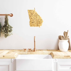 In a modern kitchen featuring a white sink and wooden countertop, the Totally Bamboo Destination Georgia Cutting Board measuring 11-3/4" x 13-1/4" adds charm to the decor. A bamboo cutting surface and gold faucet complement the look, while hanging herbs and wooden cooking utensils contribute a rustic touch to the bright, minimalist space.