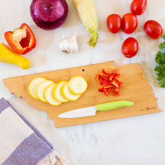 A Totally Bamboo Destination Tennessee Cutting Board, measuring 18-1/2" x 7", is elegantly shaped like the state and holds sliced yellow squash and chopped red bell pepper. Surrounding it are fresh vegetables: red onion, corn on the cob, garlic, cherry tomatoes, zucchini, and lettuce on a marble countertop with a cloth nearby.