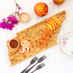 A Totally Bamboo Destination Tennessee Cutting Board, measuring 18-1/2" x 7", holds shrimp, salmon, sliced peaches, and dip. Nearby, a flower branch, whole peaches, and a small plate with forks are arranged on the white surface as part of the Destination Series.