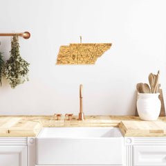 In a minimalist kitchen, a large white farmhouse sink with copper fixtures takes center stage. Just above the sink, a Destination Tennessee Cutting Board by Totally Bamboo—measuring 18-1/2" x 7"—hangs as an exquisite piece of laser-engraved artwork. Dried herbs dangle elegantly from a nearby copper bar, while wooden utensils and the bamboo cutting board rest on the counter.