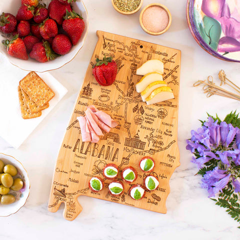 A Totally Bamboo Destination Alabama Cutting Board, measuring 10" x 15-3/4", is shaped like the state of Alabama and showcases an assortment of cheese, ham, apple slices, strawberries, and crackers. It rests on a marble countertop next to a bowl of strawberries, a dish of olives, and a bouquet of purple flowers.