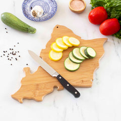 The Destination Oahu Cutting Board, 14" x 11", from Totally Bamboo, features sliced yellow and green zucchini beside a knife. Nearby, fresh tomatoes, lettuce, a zucchini, peppercorns, salt, and a blue-patterned plate with garlic are elegantly arranged on the marble surface as laser-engraved artwork adds charm.