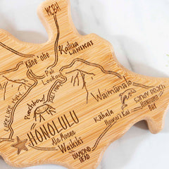 The Totally Bamboo Destination Oahu Cutting Board, measuring 14" x 11", showcases a bamboo cut-out shaped like Oahu. It features laser-engraved artwork highlighting iconic locations such as Honolulu, Waikiki, Kailua, and Sandy Beach. The detailed design includes roads and a star marking the Honolulu area, making it an exquisite bamboo cutting surface.