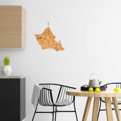 A minimalist kitchen showcases a small round table with two black chairs, featuring a teapot on a tray and three lemons. The white wall is decorated with a laser-engraved wooden map of Idaho, while the geometric vase filled with greenery pairs beautifully with the Totally Bamboo Destination Oahu Cutting Board, 14" x 11", placed on the countertop.