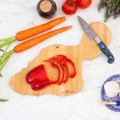 As part of Totally Bamboo's Destination Series, the Destination Maui Cutting Board, measuring 15-1/2" x 9-1/8", is uniquely designed in the shape of an island and showcases exquisite laser-engraved artwork. It elegantly supports sliced red bell peppers adjacent to a knife, while carrots, tomatoes, garlic, vinegar, and lavender are beautifully displayed on a white surface.