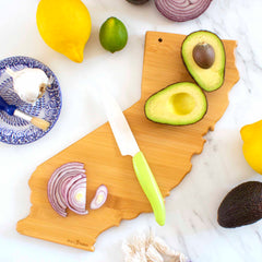 The Totally Bamboo Destination California Cutting Board, measuring 11" x 14-1/4", features a detailed laser-engraved design of the state and serves as a versatile bamboo cutting surface. It holds a knife, onion slices, and an avocado. Surrounding it are a lemon, lime, and whole avocado. Adding a touch of elegance to the setting is a blue patterned dish containing cream with an accompanying brush.