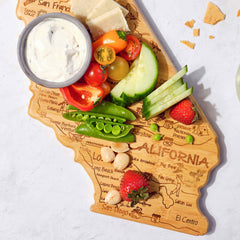 The Destination California Cutting Board by Totally Bamboo, measuring 11" x 14-1/4", presents a delightful array of cherry tomatoes, cucumber slices, snap peas, strawberries, nuts, cheese, and a dip. This uniquely shaped cutting board is embellished with laser-engraved artwork highlighting the names and landmarks of various California regions.