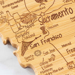 The Destination California Cutting Board from Totally Bamboo, measuring 11" x 14-/4", features a meticulously carved wooden map of Northern California. It highlights cities including Sacramento, San Francisco, Oakland, and San Jose with laser-engraved artwork. This unique bamboo cutting surface is further embellished with delightful local symbols like Napa's grapes and Santa Cruz's computer chip.