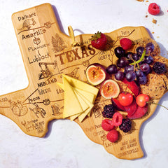 The Destination Texas Cutting Board, 14" x 13-3/8" by Totally Bamboo, is expertly engraved with city names and displays a delightful assortment of cheeses, grapes, figs, strawberries, raspberries, and crackers. Perfect as Texas kitchen decor, this board rests on a light surface with some crumbs nearby.