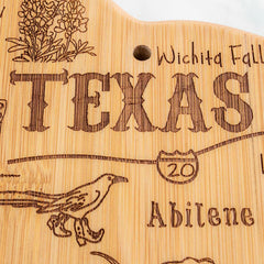 The Destination Texas Cutting Board by Totally Bamboo, measuring 14" x 13-3/8", is a stunning kitchen decor piece. This wood-crafted board features engraved images of flowers, a bird, and cities like Wichita Falls and Abilene. It even includes an illustration of a highway sign with "20," making it perfect as both a decorative item and a functional bamboo cutting board.